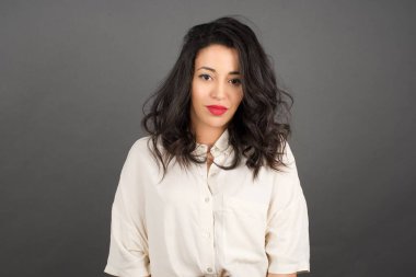 Gayri resmi giyinmiş güzel esmer kadın kameraya bakıyor. Gri arkaplanda duruyor. 