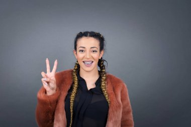 Gri duvara yaslanmış genç bir kadın iki numarayı gösterip işaret ederken kendinden emin ve mutlu bir şekilde gülümsüyor..