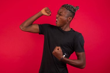 Kırmızı arka planda siyah tişört giyen çekici Afro-Amerikalı bir adam zaferi kutluyor yumruklarıyla havaya yumruk atıyor ve parlak dişli bir gülümsemeyle.