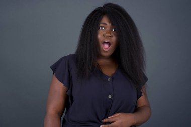 Etkileyici yüz ifadeleri. Şaşırmış Afrikalı Amerikalı kadın çenesini kapalı tutuyor, çizgili süveter giyiyor, boş gri duvarda gördüklerinden sersemlemiş hissediyor.