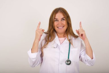 Medikal üniforma giyen, her iki işaret parmağıyla işaret eden başarılı, dost canlısı görünümlü, olgun, beyaz doktor kadının yatay çekimi, boş bir fotokopi duvarında şok edici bir şey olduğunu gösteriyor..