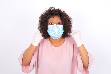 Yüz maskesi takan kadın ve beyaz stüdyo arka planında izole edilmiş yumrukları olan günlük kıyafetler.