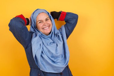 Güven ve cinsel ilişki konsepti. Orta yaşlı tesettürlü çekici müslüman kadın portresi, kendine güvenen bir ifadeyle gülümseyerek ellerini başının üstüne koyuyor. Arkaplanda duruyor.