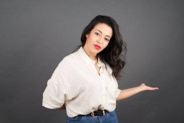 Güzel esmer kadın avuçlarını açık tutarak gündelik giyinmiş. Gri arkaplanda duruyor. 