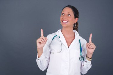 Gri arka planda parlak makyajlı başarılı, dost canlısı genç doktor kadının yatay görüntüsü heyecanlı bir şekilde işaret ederek boş fotokopi duvarında şok edici bir şey olduğunu gösteriyor..