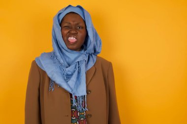 Vücut dili. Sarı duvara karşı poz veren, suratını asan, bir şeye karşı hoşnutsuzluk gösteren güzel Afro-Amerikan Müslüman kadın..
