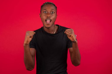 Siyah tişörtlü, kırmızı arkaplanlı güçlü, kararlı Afro-Amerikan bir adamın belden yukarı portresi. Kollarını sıkarak, sevinç ve heyecanla bağırıyor. Zafer, başarı ve başarı kavramı.