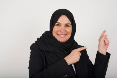 Reklam konsepti. Gri arkaplandaki siyah tesettürlü güzel Müslüman kadın işaret parmağıyla işaret ediyor ve boş kopya alanına bakıyor..