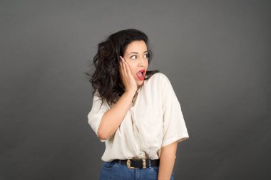 Gayri resmi giyinmiş güzel esmer kadının kafası karışmış. Gri arkaplanda duruyor. 