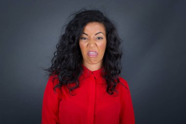 Kıvırcık saçlı, gri gömlekli iğrenç esmer kadın gri duvara poz veriyor, kaşlarını çatıyor, bir şeyden tiksindiğini gösteriyor..