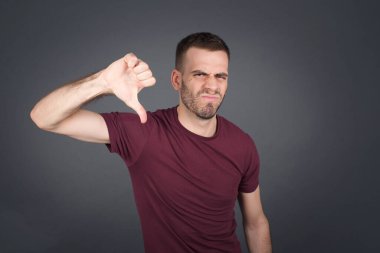 Memnuniyetsiz Avrupalı bir adam hoşnutsuzluk işareti gösterir, baş parmağını aşağıda tutar, hoşlanmadığını ifade eder, hoşnutsuzca kaşlarını çatar, gri arka plan üzerine. Vücut dili kavramı.