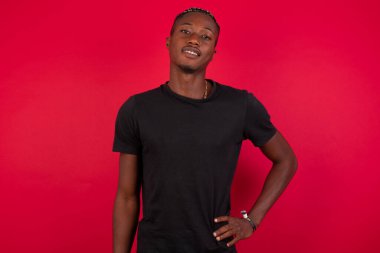 Kırmızı arka planda siyah tişört giyen neşeli Afro-Amerikalı adamın stüdyo fotoğrafı elini kalçasında tutuyor, geniş bir şekilde gülümsüyor, arkadaşlarının doğum gününde hazırladığı sürprizi bekliyor.