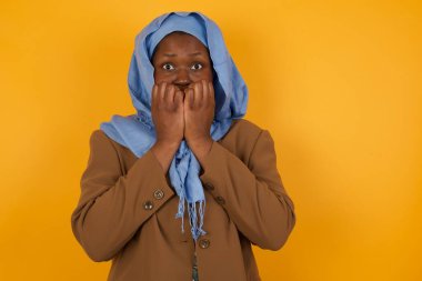 Endişe. Güzel bir Afrikalı Amerikalı müslüman kadının kavramsal görüntüsü. Ağzını elleriyle kapatıyor ve sarı arka planda duruyor. Bir şeyden ya da birinin çivi yemesinden korktun mu?.