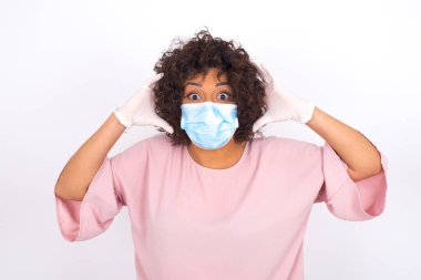  Yüz maskesi takan kafası karışık bir kadın ve beyaz stüdyo arka planında izole edilmiş günlük kıyafetler.