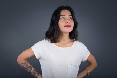 Neşeli kızın belinden yukarı bakıp bir şeyler düşündüğü bir sahne. Ellerini kalçaya koy, yüz ifadenden şüphe et. Gri duvara yaslanmış bir şey olup olmadığını merak ediyorum..