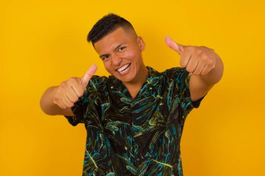 Tecrit edilmiş arka planda yaşayan İspanyol genç adam elleriyle olumlu jest yapmayı onaylıyor, gülümsüyor ve başarı için seviniyor. Kameraya bak, kazanan jest..