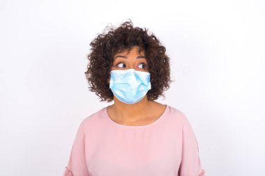 Yüz maskesi takan ve sıradan giyinen bir kadın beyaz stüdyo arka planında izole edilmiş bir şekilde yüzünü çeviriyor.