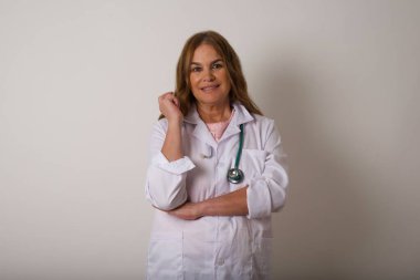 Tıbbi üniforma giyen neşeli, olgun, beyaz bir kadın resmi. Yüzüne yakın eli gri bir duvarın önünde duruyor. İyi haberleri dinledikten sonra kameraya mutlu bir ifadeyle bakmak. Kendine güvenen kız..