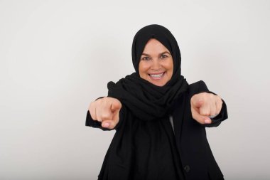 Siyah tesettürlü, iki parmağıyla kamerayı işaret eden, ağzı açık, arka planda izole bir şekilde yukarı bakan, şaşırmış, genç Müslüman kadının yakın plan portresi: