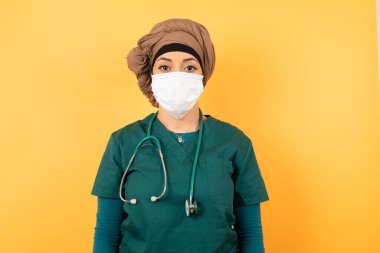 Yeşil üniformalı güzel Müslüman kadın kameraya bakıp bir şey düşünürken belini kaldır. İki kolu aşağıda, tarafsız yüz ifadesi bir şeyler olduğunu merak ediyor..