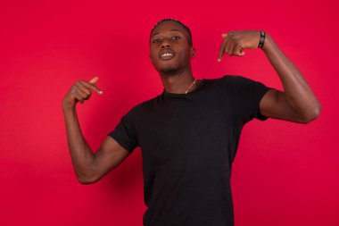 Kırmızı arka planda siyah tişört giyen, neşeli bir şekilde gülümseyen ve olumlu duygular ifade eden Afro-Amerikan yakışıklı bir adam. Çocuk çok antrenman yaptı, o da kaslarını göstermek istiyor.