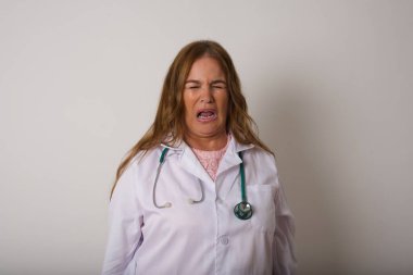 Negatif insan ifadeleri ve tavırları. Kızgın, tatminsiz, beyaz doktor kadın, tıbbi üniforma giyen, çirkin bir ifade takınıyor, kaşlarını çatıyor, beyaz arka planda soyutlanıyor. Güvensizlik ve nefret.