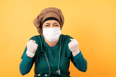 Arap kadın doktor portresi, yüz maskesi takmış, sağlık üniforması giymiş, yumruklarını sıkmış, sevinçle ve heyecanla hedefe sesleniyor.. 