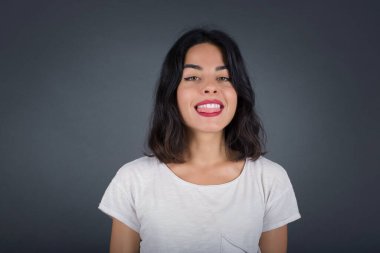 Mutlu ve komik yüzlü, gülümseyen ve dil gösteren güzel beyaz bir kadın. Günlük giysiler giyip gri stüdyonun arka planında dikiliyorlar.. 