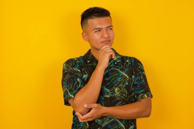 Düşünceli, düşünceli genç bir adam elini çenesinin altında tutuyor, düşünceli bir şekilde yukarı bakıyor, gündelik kıyafetler giyiyor, sarı arka planda duruyor ve mesajın için boş yer var.