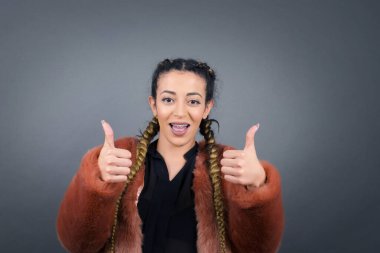 Gri arka planda izole edilmiş, olumlu içerik ve mutluluk duygularıyla baş parmaklarını gösteren iyimser genç bayana yakın çekim.