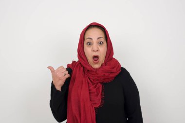 Şok olmuş olgun Müslüman kadının başparmağıyla işaret ettiği yatay çekim beyaz arka planda izole edilmiş boş bir duvara işaret ediyor. Şuna bir bak.. 