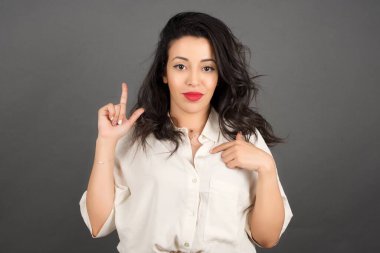 Güzel esmer kadın, sıradan giyinmiş, işaret ediyor. Gri arkaplanda duruyor.  