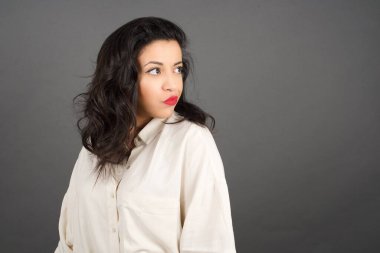 Güzel esmer kadın, sıradan giyinmiş, gözlerini kaçırıyor. Gri arkaplanda duruyor.  