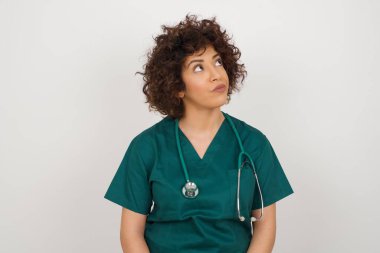 Şaşkın, güzel, Arap doktor kadın tıbbi üniforma giyiyor, kaşlarını çatıyor, yukarı bakıyor.