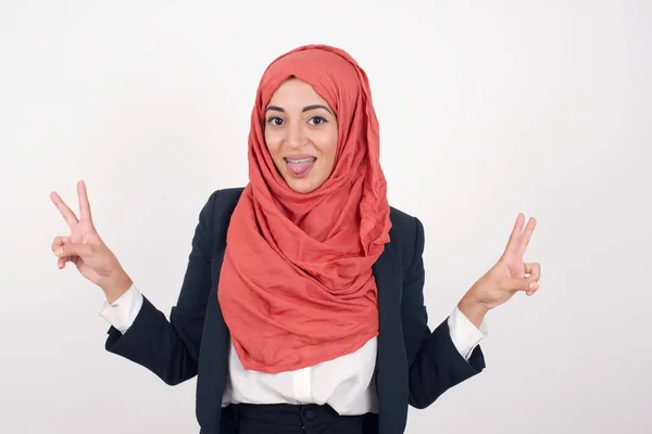 美しいイスラム教徒の女性の屋内肖像画は 楽観的な笑顔で灰色の背景に隔離された黒いブレザーとヒジャブを着用し 両手で平和や勝利のジェスチャーを示し 友好的に見えます Vサイン — ストック写真
