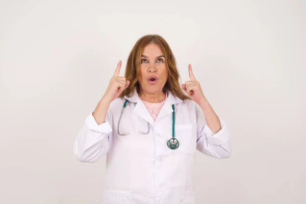 美丽而成熟的高加索医生的画像 身穿医疗制服的女人在与世隔绝的背景下惊讶地看着 用手指指出一些奇怪的东西 — 图库照片