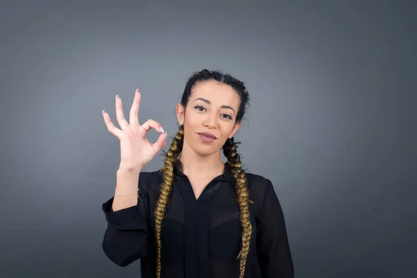 Glad attractive woman shows ok sign with hand as expresses approval, has cheerful expression, being optimistic. Standing against white wall.