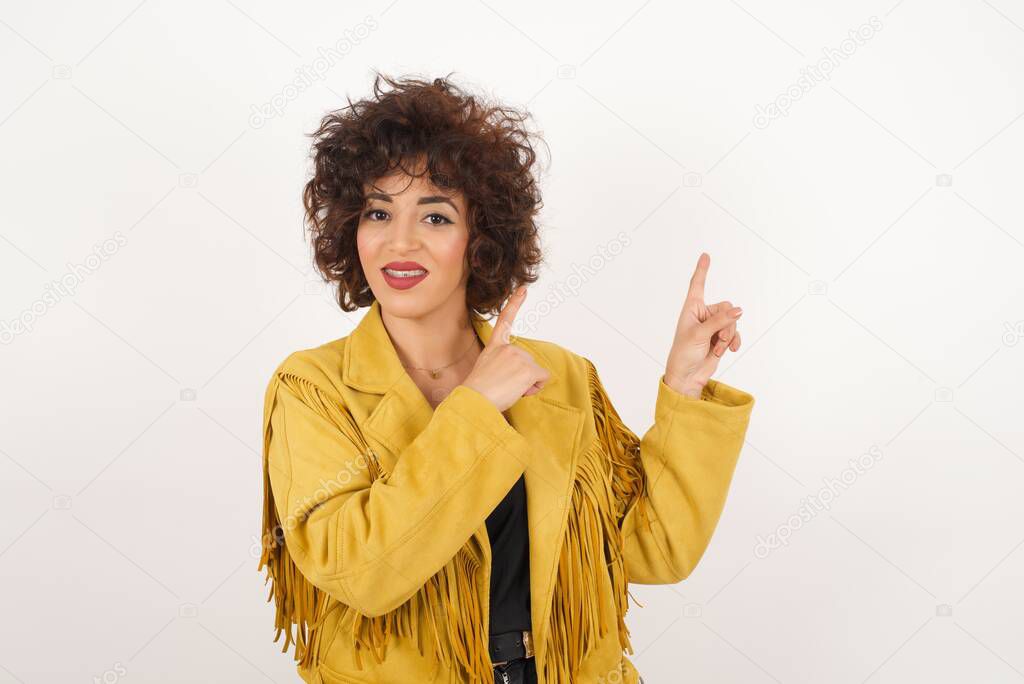 Advertisement concept. Positive, pretty, woman with beaming smile in jeans shirt on grey background is pointing with her two index finger and looking on empty copy space.