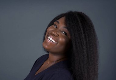 Utangaç kız kameraya bakarak gülümsüyor. Gri stüdyo arka planına zımparalayan Afrikalı Amerikalı kadın utangaç hissediyor.