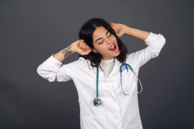 Güven ve cinsel ilişki konsepti. Çekici genç bir doktorun portresi. Kendine güvenen bir ifadeyle gülümsüyor ve ellerini başının üstünde tutuyor. Gri arkaplanda duruyor.