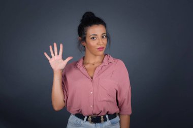 Gri duvara yaslanmış genç bir kadın, mutlu ve kendinden emin gülümserken beş numarayı gösterip işaret ediyor..