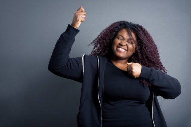 Çekici Afro-Amerikalı kadın gündelik giysiler içinde zaferi kutluyor yumruklarıyla havaya yumruk atıyor ve gri bir stüdyonun arka planında ışıldayan dişli bir gülümsemeyle fotokopi çekiyordu.