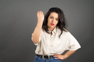 Güzel esmer kadın, gelişigüzel giyinmiş. Gri arkaplanda duruyor. 