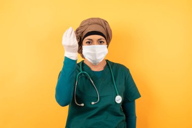 What the hell are you talking about. Shot of frustrated Young beautiful Muslim doctor woman in green medical uniform gesturing with raised hand doing Italian gesture, frowning, being displeased and confused with dumb question.