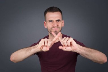 Gri duvara yaslanmış, izole edilmiş arka plan üzerinde sıradan kıyafetler giyen genç yakışıklı bir adam. Reddedilme ifadesi negatif işaret yaparak parmaklarını çapraz yapıyor mu?.