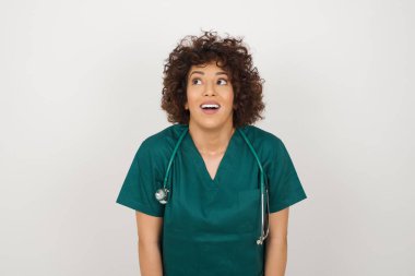 İnsanlar ve ani tepki kavramı. Şaşkın Arap doktor kadın tıbbi üniforma giyiyor, omuz silkiyor, yana bakıyor, arkadaşlarıyla yeni planları için mutlu ve heyecanlı. Gri duvarda izole edilmiş..