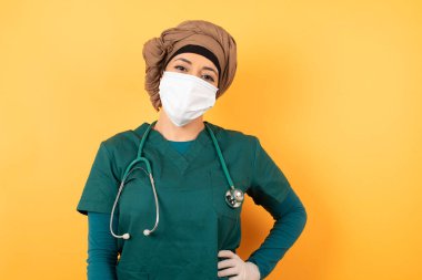 Yüz maskeli Arap kadının stüdyo fotoğrafı. Elini kalçasında tutuyor, geniş bir şekilde gülümsüyor. Sürpriz olmasını bekliyor.