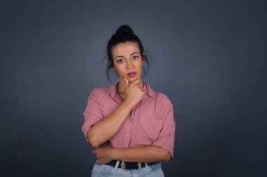 Düşünceli gülümseyen kızın portresi elini çenesinin altında tutuyor, doğrudan kameraya bakıyor, ilgi çekici bir şey dinliyor, sıradan giyiniyor, gri duvara karşı poz veriyor..