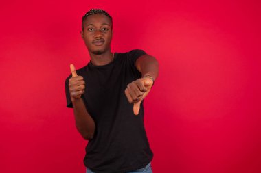 Kırmızı arka plan üzerinde siyah tişört giyen genç ve dalgın bir Afro-Amerikan adam iyiye-kötüye işaret yapıyor. Keyifsiz ve etkilenmemiş sıradan kıyafetler giyip gri duvarın önünde dikiliyor..