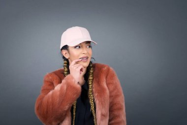 Hayalperest bir kadın, düşünceli bir ifadeyle, kameraya bakıyor, elini yüzüne yakın tutuyor, parmağını ısırıyor ve hoş bir şey düşünüyor, gri duvara karşı poz veriyor..